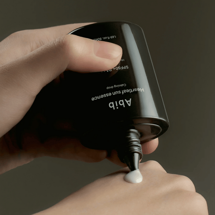 Hand som klämmer ut Abib Heartleaf Sun Essence Calming Drop på ovansidan av en annan hand, med mörk bakgrund och tydlig visning av produktens konsistens.