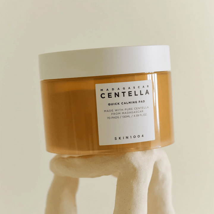 Burk med SKIN1004 Madagascar Centella Quick Calming Pad placerad på en skulpturliknande vit piedestal – bärnstensfärgad behållare med vit etikett och lock.
