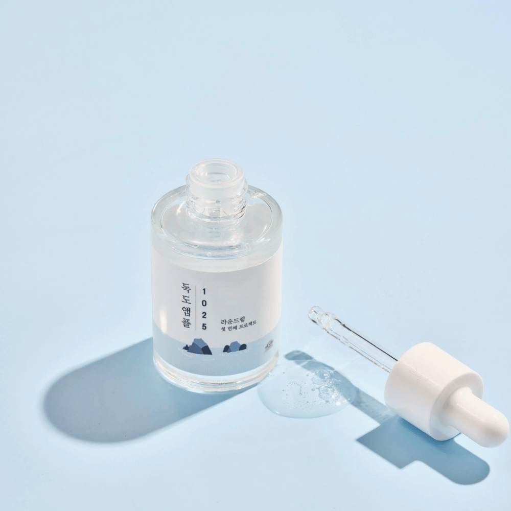 Uma garrafa aberta de 1025 Dokdo Ampoule com um conta-gotas ao lado, contra um fundo azul que cria uma sombra suave.