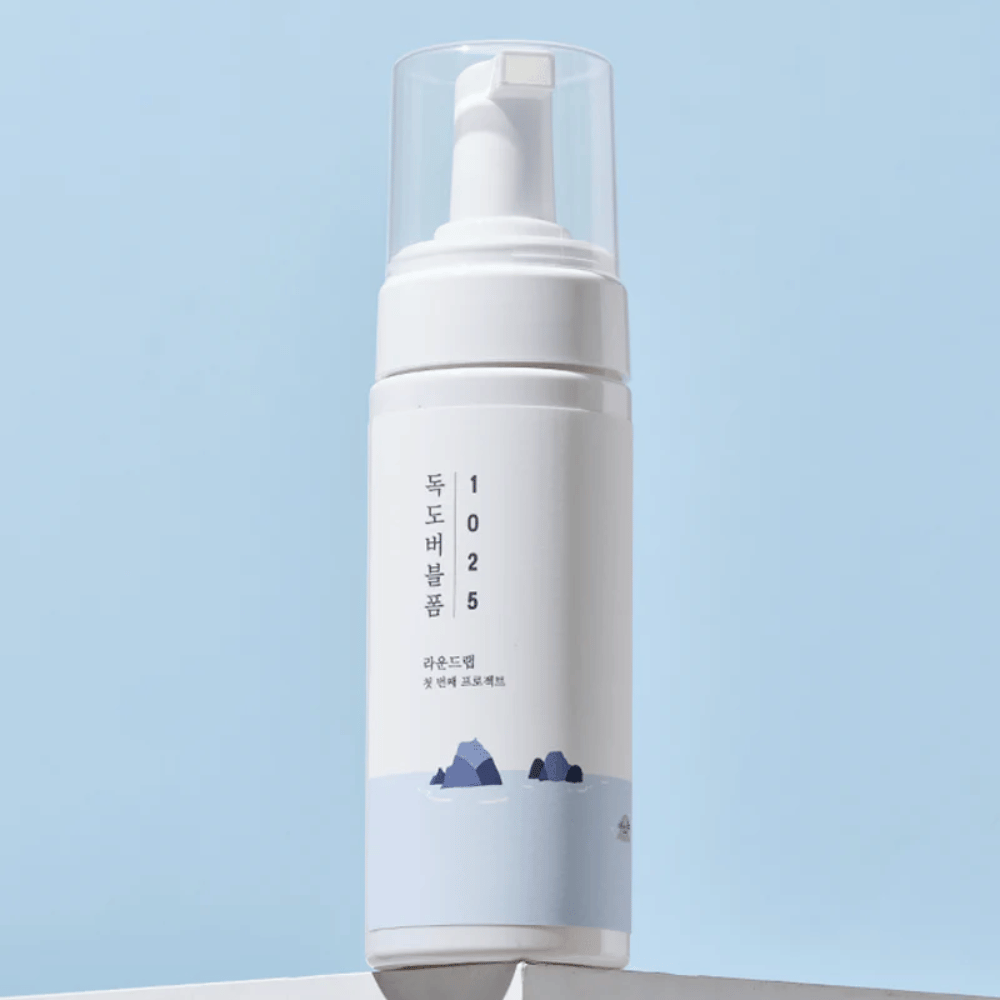  O produto é um "1025 Dokdo Bubble Foam" da ROUND LAB, embalado em um frasco de bomba branco com design minimalista. É destinado à limpeza profunda do rosto sem irritação.