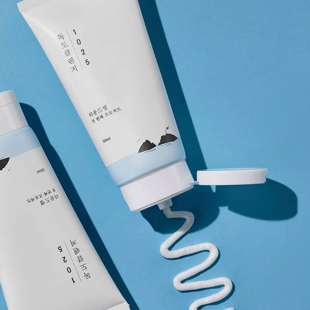 Dois tubos de 1025 Dokdo Cleanser em fundo azul, um com tampa aberta e uma linha ondulada de limpeza espremida.