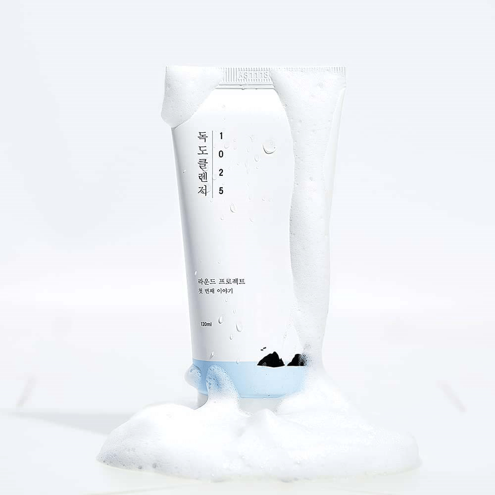 1025 Dokdo Cleanser em tubo branco coberto de espuma, com gotas de água e espuma ao redor da base, isolado sobre fundo claro.