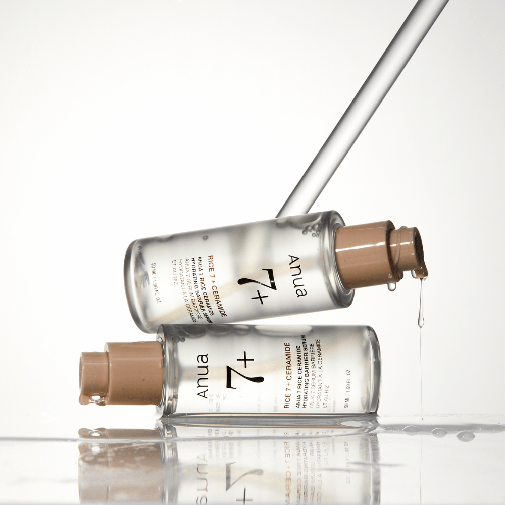 A imagem mostra duas garrafas transparentes de "Anua 77 Rice + Ceramide Hydrating Barrier Serum" com gotas escorrendo de uma pipeta. O sérum, que é enriquecido com arroz e ceramidas, é destinado a hidratar e fortalecer a barreira da pele. As garrafas são elegantemente desenhadas, o que reforça a sensação premium do produto.