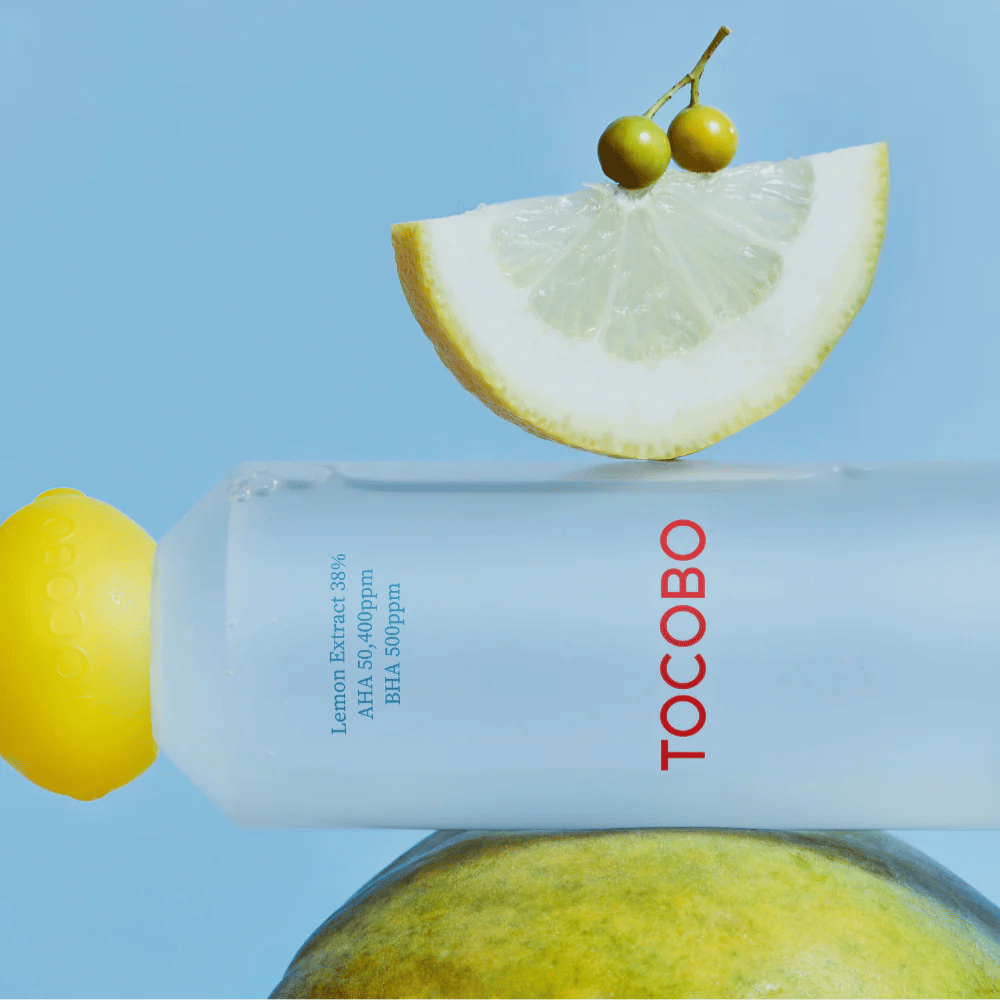 Garrafa com TOCOBO AHA BHA Lemon Toner, decorada com uma tampa em forma de limão. Uma fatia de limão e pequenas bagas verdes equilibram-se em cima da garrafa contra um fundo azul.