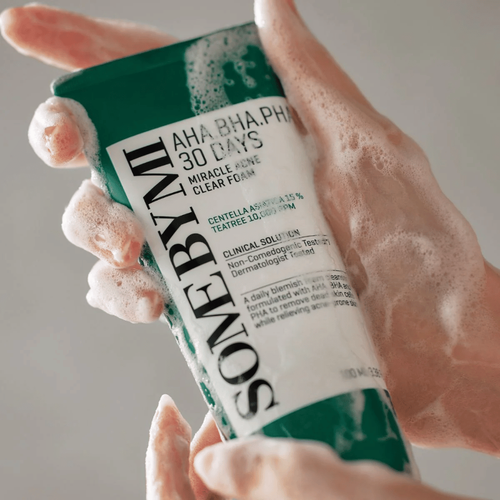 Uma pessoa segura um tubo de "SOME BY MI AHA, BHA, PHA 30 Days Miracle Acne Clear Foam" com conteúdo espumante sobre as mãos. O produto é destinado ao uso diário para combater a acne e limpar os poros com seus ingredientes ativos como centella, óleo de tea tree e ácidos como AHA, BHA e PHA.