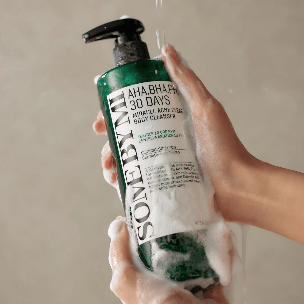 Uma pessoa segura um frasco SOME BY MI AHA.BHA.PHA 30 Days Miracle Acne Clear Body Cleanser. O frasco é verde com um rótulo branco e está coberto de espuma do produto de limpeza. A pessoa lava-se com o produto enquanto a água escorre pelas mãos.