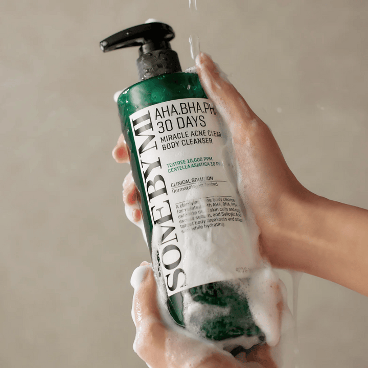 En person håller i en flaska SOME BY MI AHA.BHA.PHA 30 Days Miracle Acne Clear Body Cleanser. Flaskan är grön med en vit etikett och är täckt av lödder från rengöringsmedlet. Personen tvättar sig med produkten medan vatten rinner över händerna.