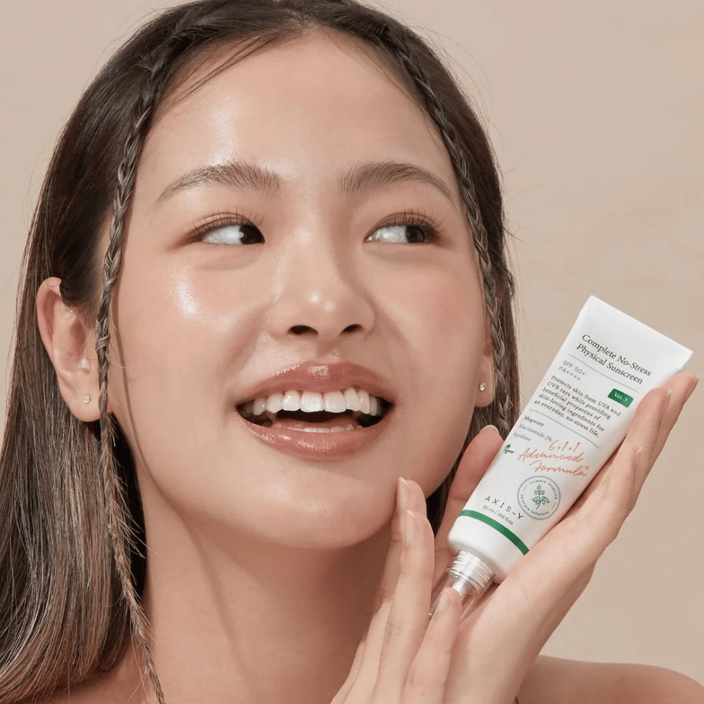 A imagem mostra uma jovem mulher sorrindo alegremente enquanto segura um tubo de AXIS-Y Complete No-Stress Physical Sunscreen. Ela tem uma aparência radiante e natural com maquiagem mínima, o que destaca o produto que ela apresenta.