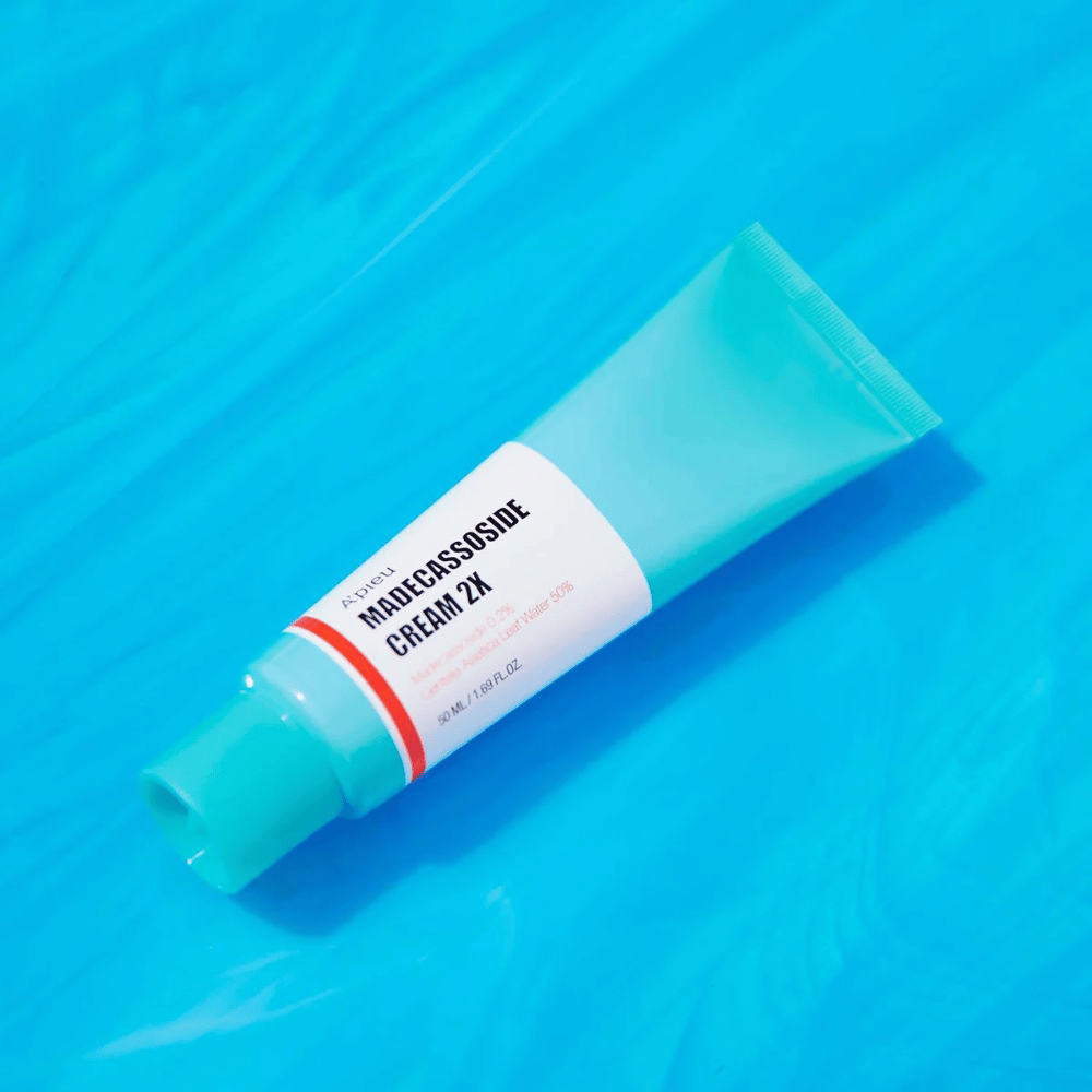 Um tubo A'PIEU Madecassoside Cream 2X está colocado diagonalmente sobre um fundo azul com rugas semelhantes à água. O tubo tem um corpo branco com uma tampa turquesa clara e um rótulo de cor vermelha a laranja onde o nome do produto e o ingrediente ativo Madecassoside 2% estão claramente impressos. O rótulo também indica a quantidade que é 50 ml / 1.8 fl oz. A apresentação clara e colorida dá uma sensação fresca e energética.