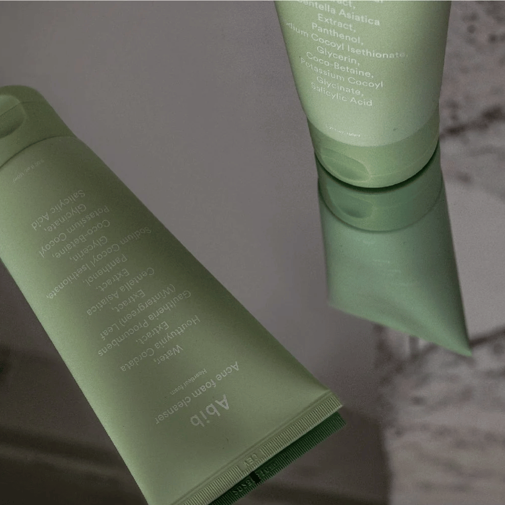 Tubo verde de Abib Acne Foam Cleanser Heartleaf deitado em uma mesa espelhada com a frente do produto e o texto refletido. Ingredientes como ácido salicílico, centella e extrato de folha de coração são claramente visíveis na embalagem.
