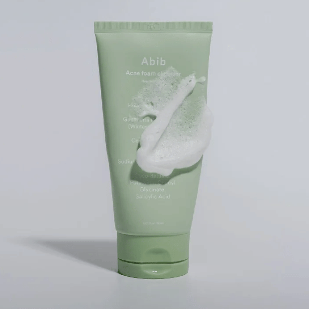Tubo verde com Abib Acne Foam Cleanser Heartleaf, coberto por uma espuma branca que ilustra o efeito de limpeza espumante. O produto contém extrato de folha de coração e ácido salicílico para pele propensa a acne.