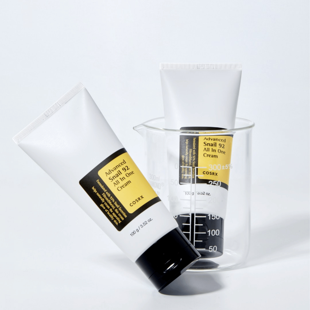 Um tubo de COSRX Advanced Snail 92 All in One Cream deitado ao lado de um cilindro de medição transparente invertido sobre um fundo branco.