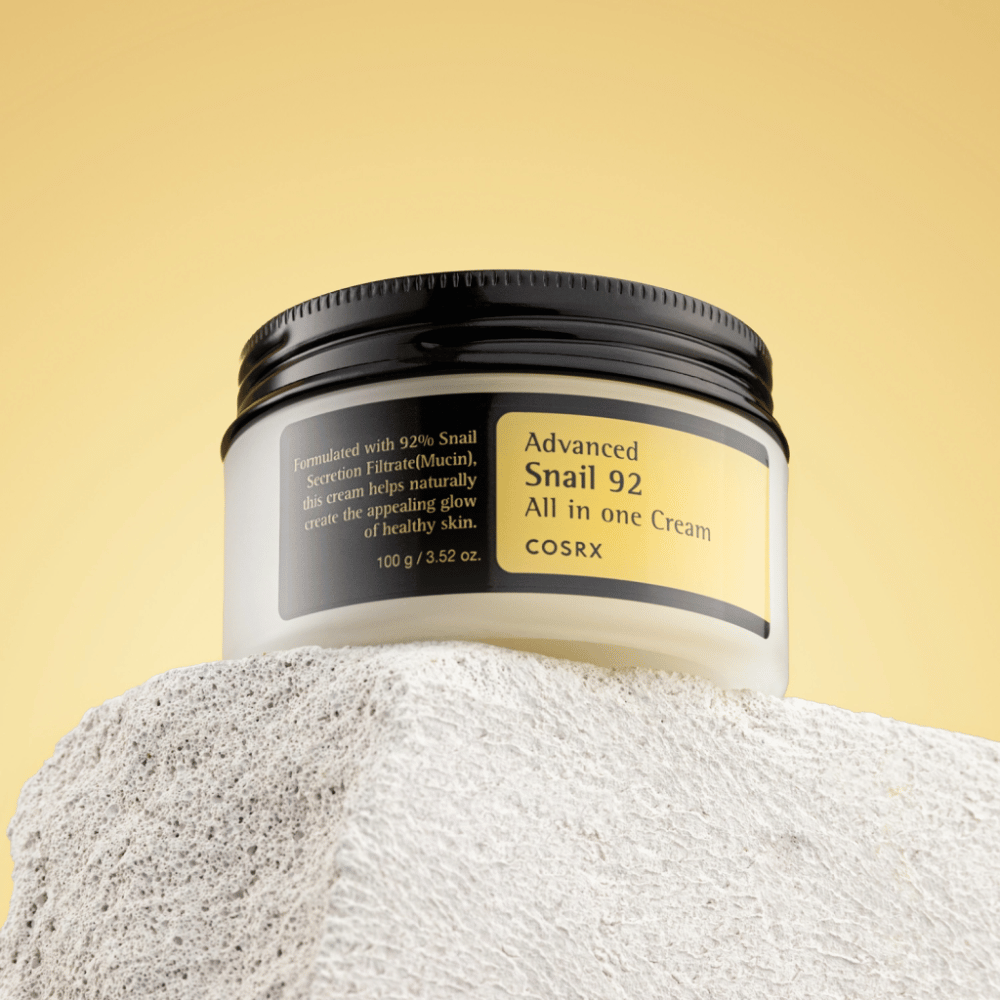 Um frasco de COSRX Advanced Snail 92 All in one Cream colocado sobre uma superfície rochosa com um fundo amarelo.