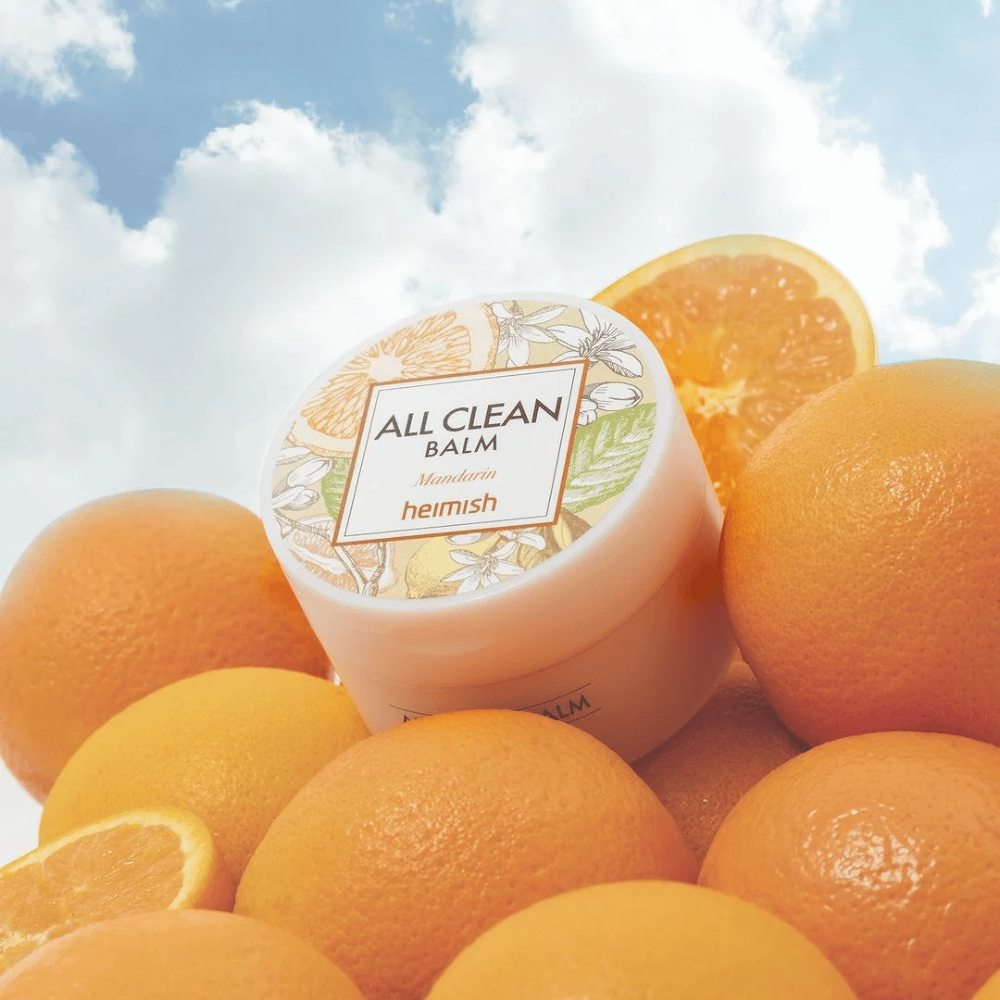 Frasco de Heimish All Clean Balm Mandarin colocado sobre uma pilha de laranjas, com um céu azul ao fundo.