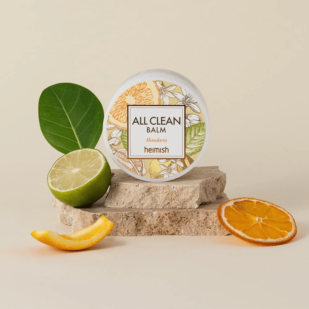 Frasco de Heimish All Clean Balm Mandarin colocado sobre pedras, rodeado por cascas de tangerina e folhas de citrinos, em frente a um fundo claro.