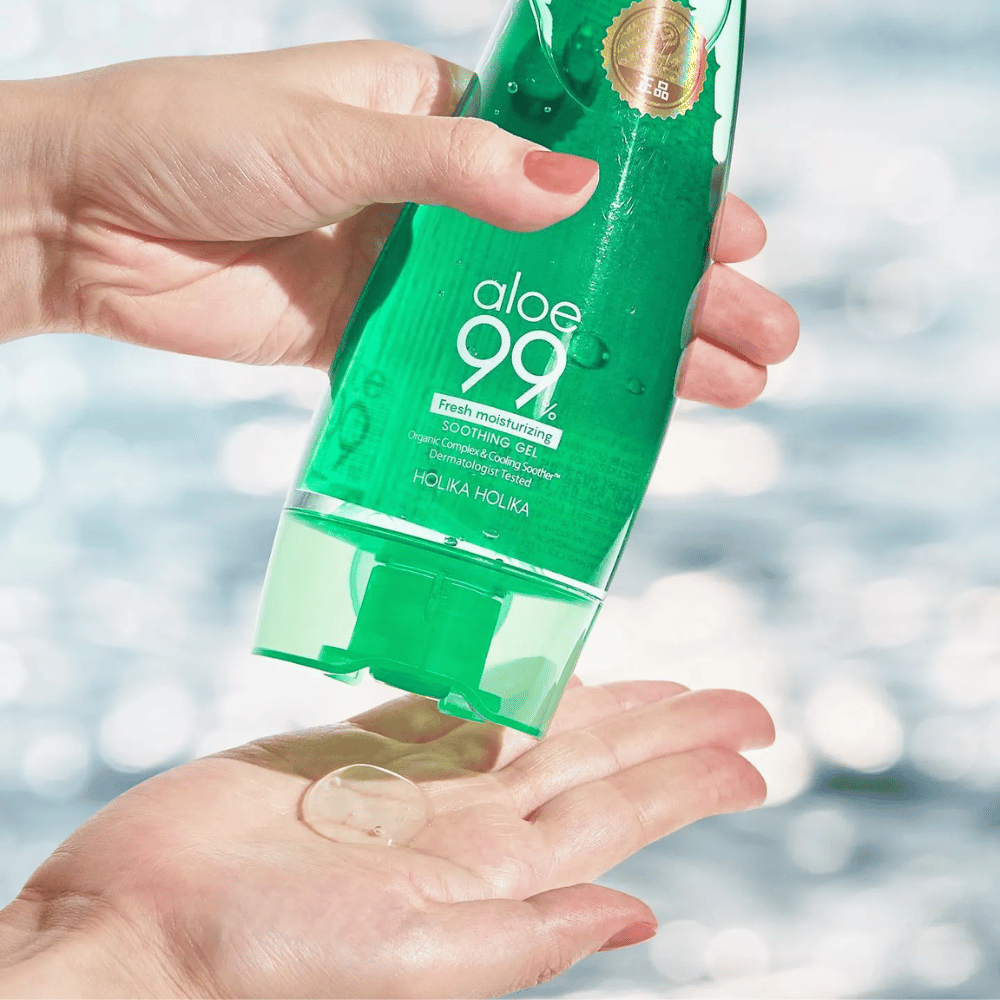 Uma mão segura um tubo verde com Holika Holika Aloe 99% Soothing Gel, enquanto outra mão captura uma porção de gel que é dispensada. O fundo está desfocado e mostra um padrão aquoso e cintilante.