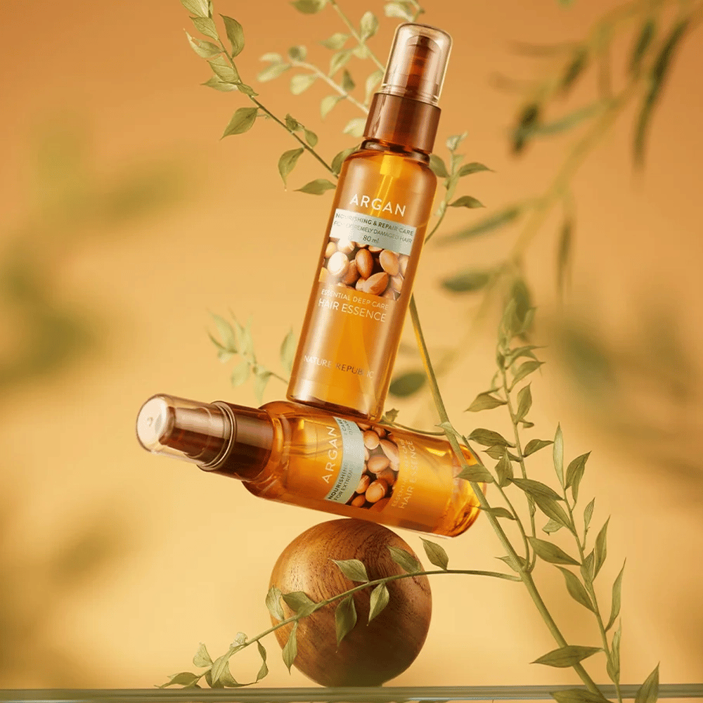 Duas garrafas Nature Republic Argan Essential Moisture Hair Oil colocadas entre folhas verdes – brilho sedoso para cabelo seco.