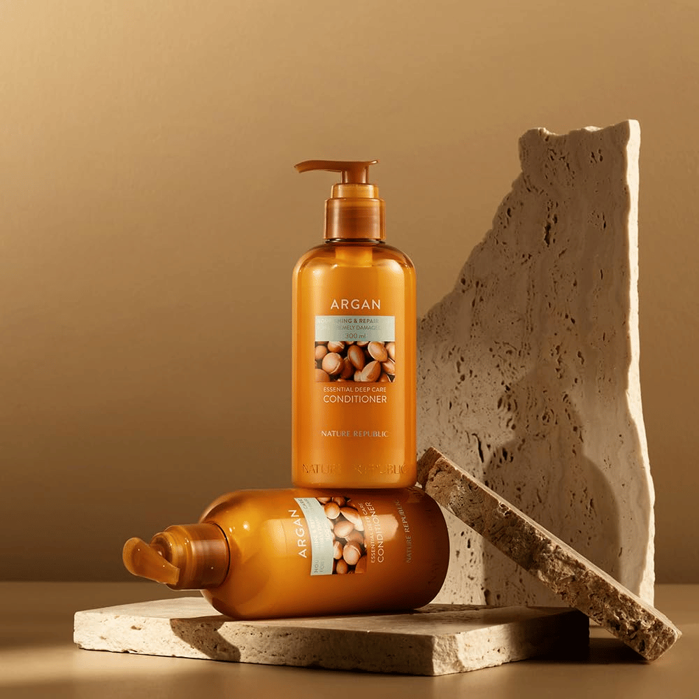 Duas garrafas de Nature Republic Argan Essential Moisture Repairing Conditioner em superfície de pedra em tom quente.