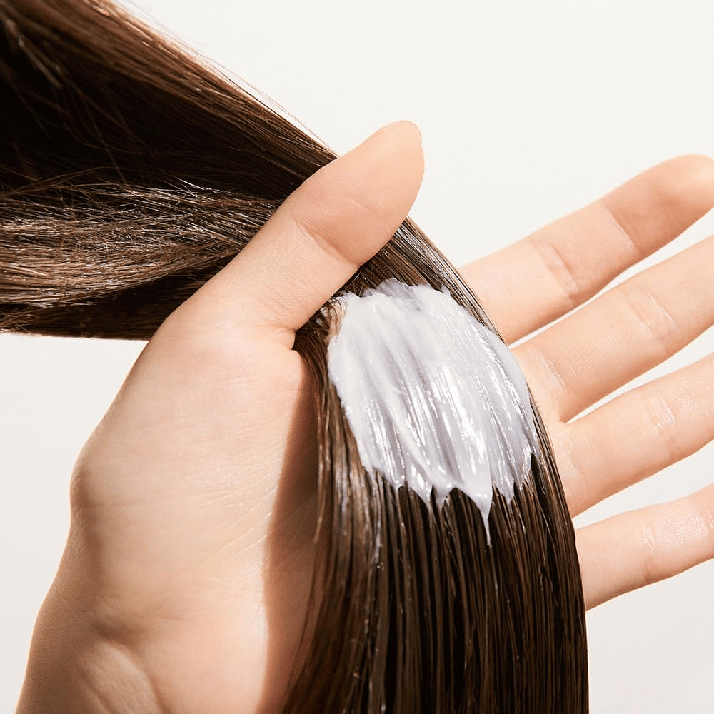 Mão aplica Nature Republic Argan Essential Moisture Repairing Hair Mask em cabelo molhado – máscara hidratante que fortalece e suaviza o cabelo.