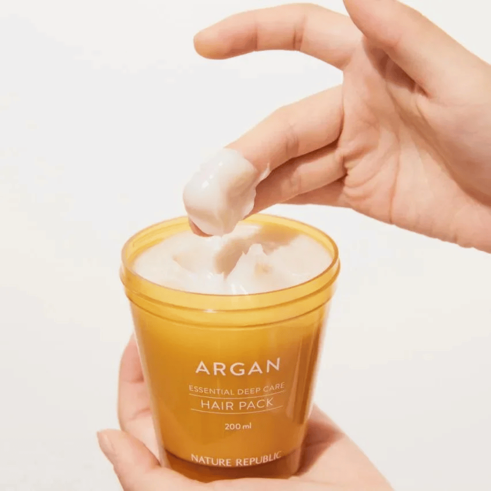 Mão pega a fórmula cremosa de Nature Republic Argan Essential Moisture Repairing Hair Mask – nutrição intensa para cabelo seco.