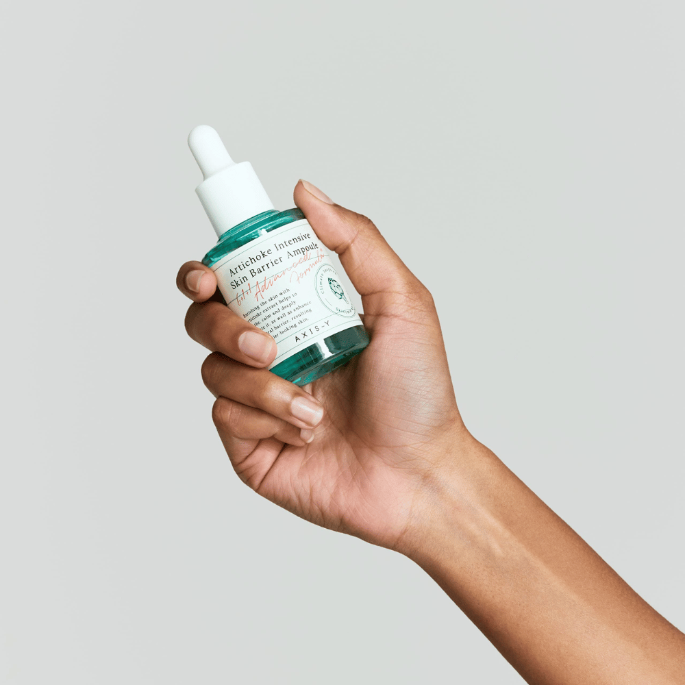 Uma mão segura uma garrafa de sérum para cuidados com a pele com a etiqueta "Artichoke Intensive Skin Barrier Ampoule.