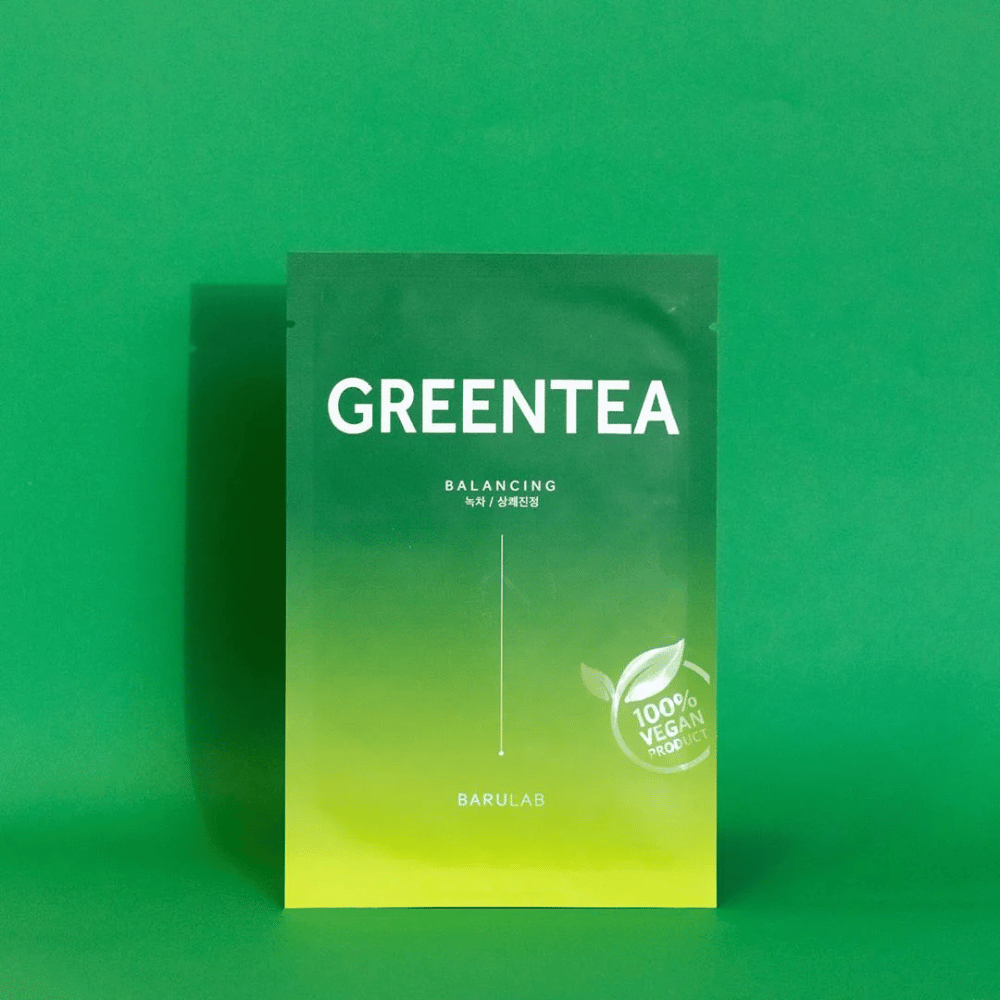 Uma embalagem de BARULAB para uma máscara "GREENTEA Balancing", marcada como "100% Vegan Product". A cor da embalagem transita do verde escuro no topo para um verde mais claro na parte inferior, contra um fundo verde uniforme.