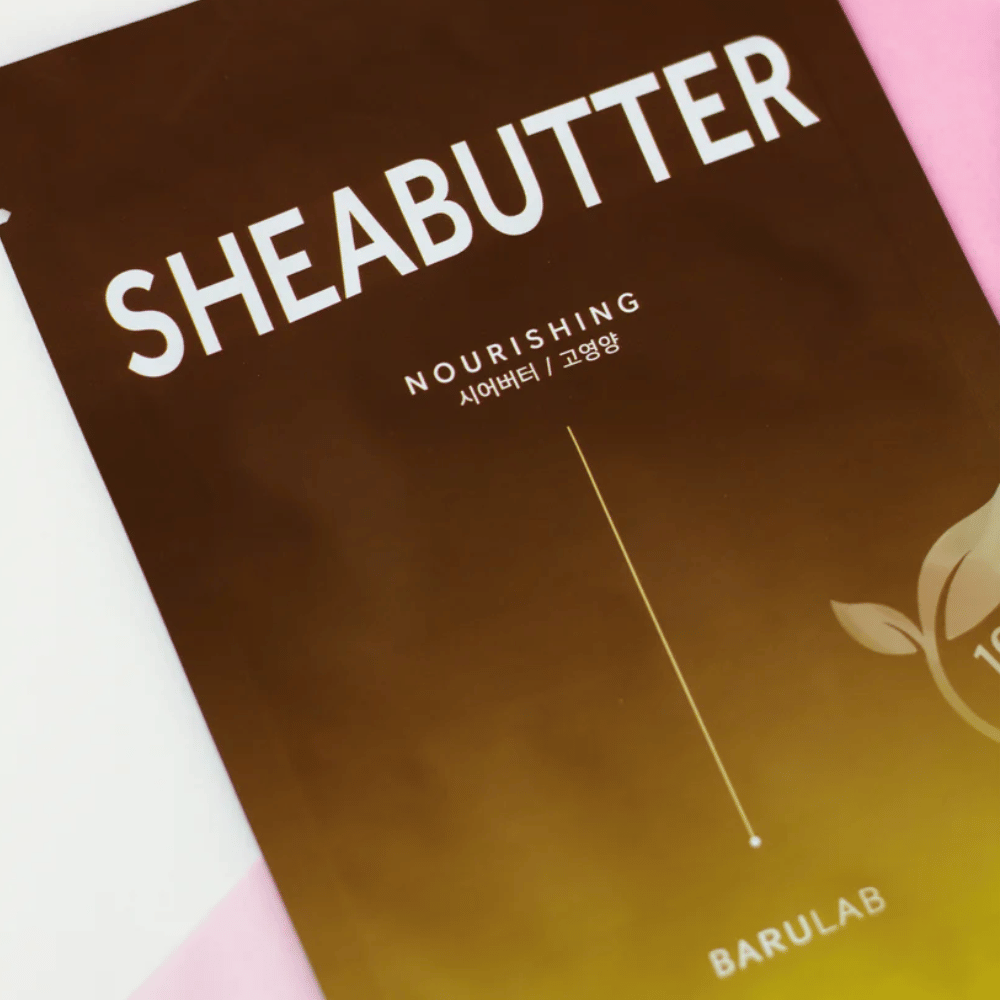 Uma imagem de close-up de uma embalagem da máscara "SHEABUTTER Nourishing" BARULAB. O produto é marcado como "100% Vegan Product" e tem um gradiente de cor de castanho escuro para castanho claro.