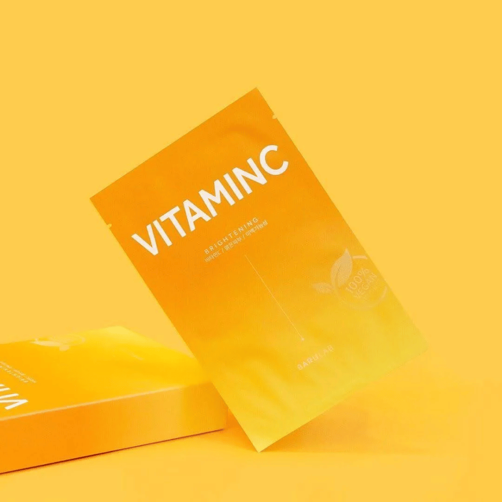 Uma pilha de embalagens de máscaras "VITAMINC Brightening" BARULAB, todas marcadas como "Produto 100% Vegano". As embalagens têm uma tonalidade em diferentes nuances de laranja e amarelo, colocadas contra um fundo amarelo uniforme.
