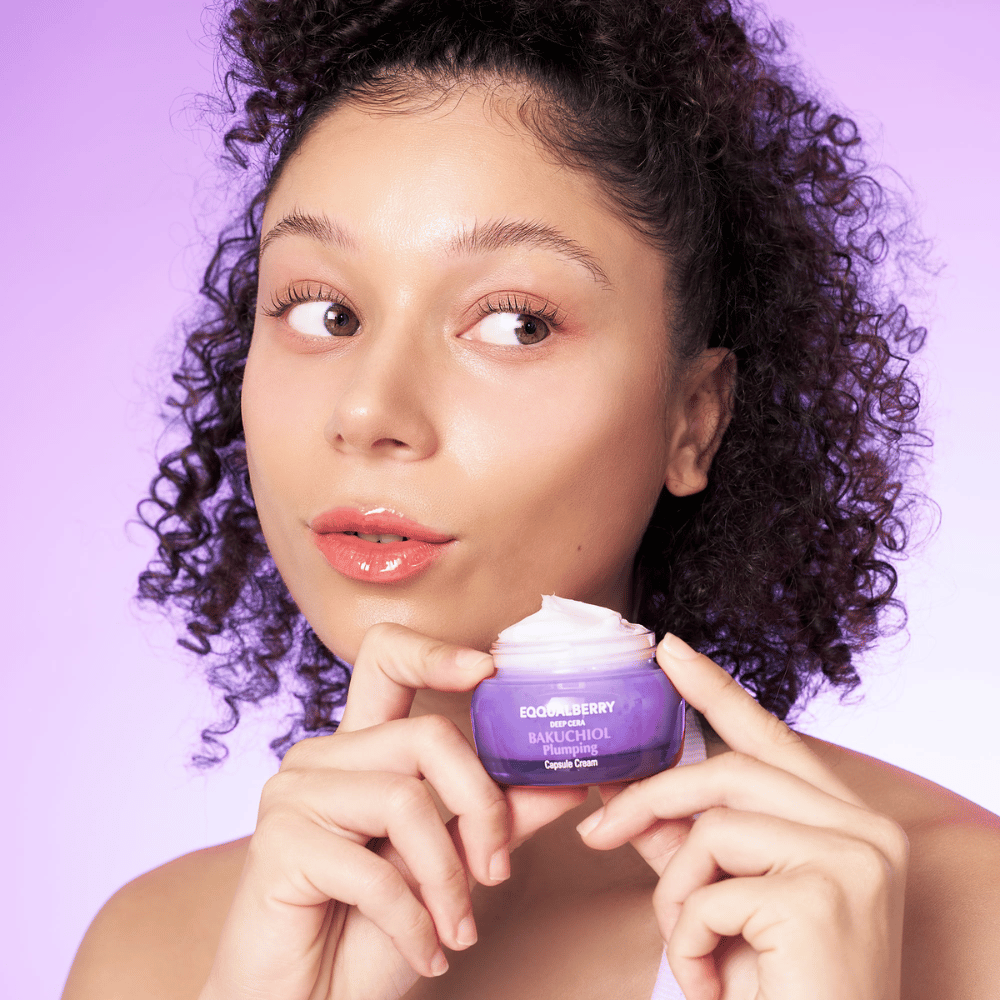 Mulher segura EQQUALBERRY Bakuchiol Plumping Capsule Cream na mão e exibe sua pele hidratada e radiante contra um fundo roxo.