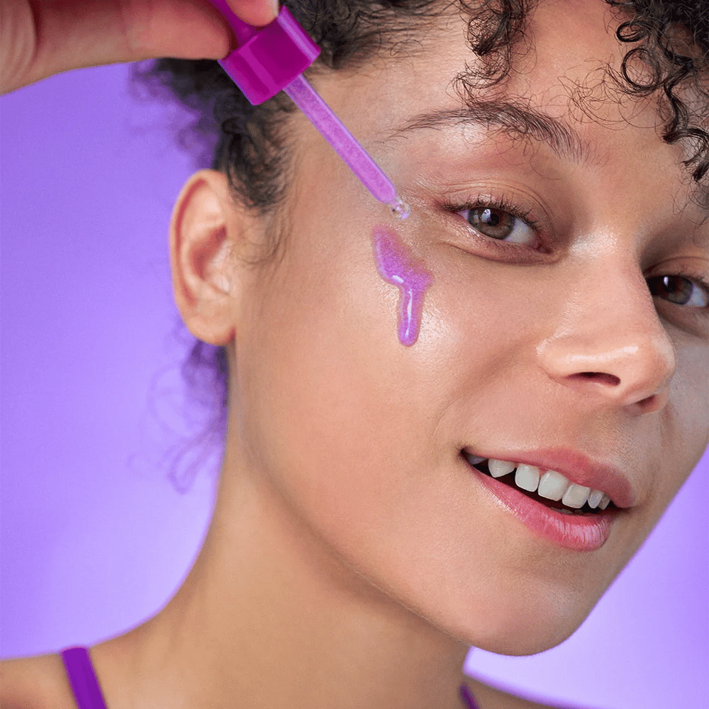 Mulher sorridente aplica um sérum cintilante roxo na pele com uma pipeta. Pele radiante e fundo roxo que combina com o tom do produto.