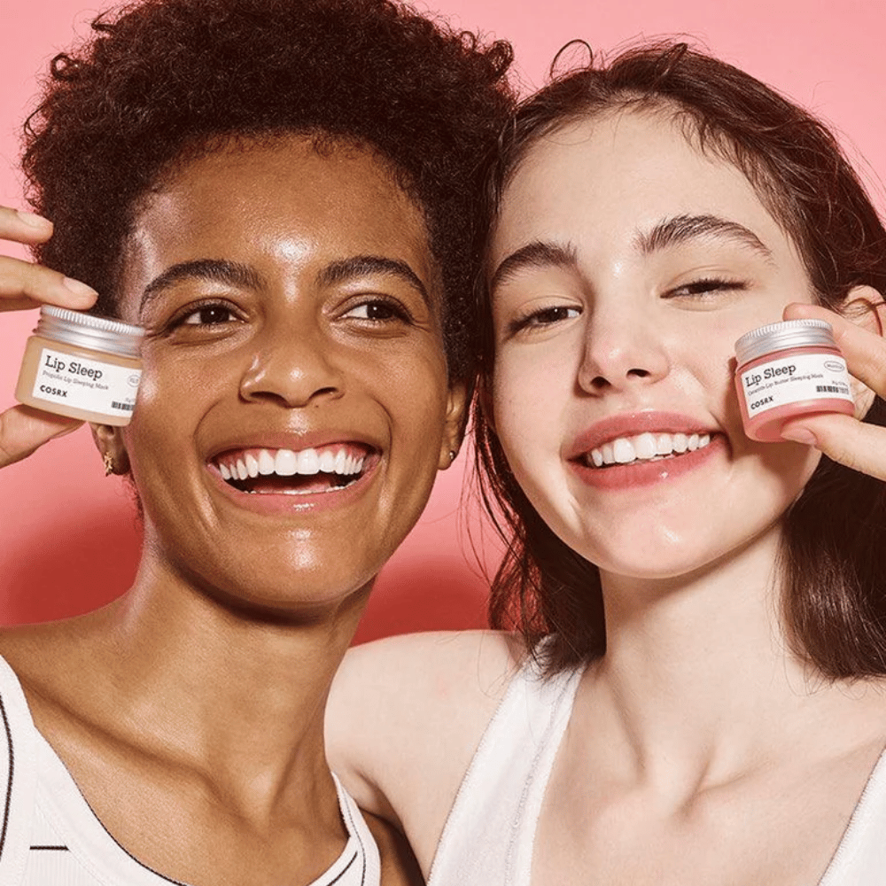 Duas pessoas sorriem e seguram potes de COSRX Lip Sleep. A imagem transmite lábios macios, hidratados e o efeito natural de brilho do produto.
