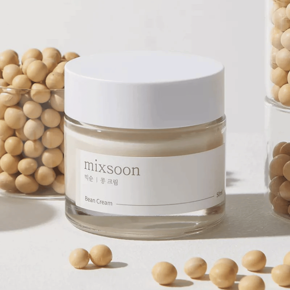 Um frasco transparente com rótulo e tampa brancos, etiquetado "mixsoon 팥 크림" e "Bean Cream" com um volume indicado de "50ml". O frasco contém um creme amarelo claro e está colocado sobre uma superfície clara. Ao lado do frasco há um recipiente de vidro cheio de grãos castanhos claros que foram derramados na superfície, sugerindo que o creme contém extrato de grão. O fundo e a iluminação proporcionam uma atmosfera quente e natural.
