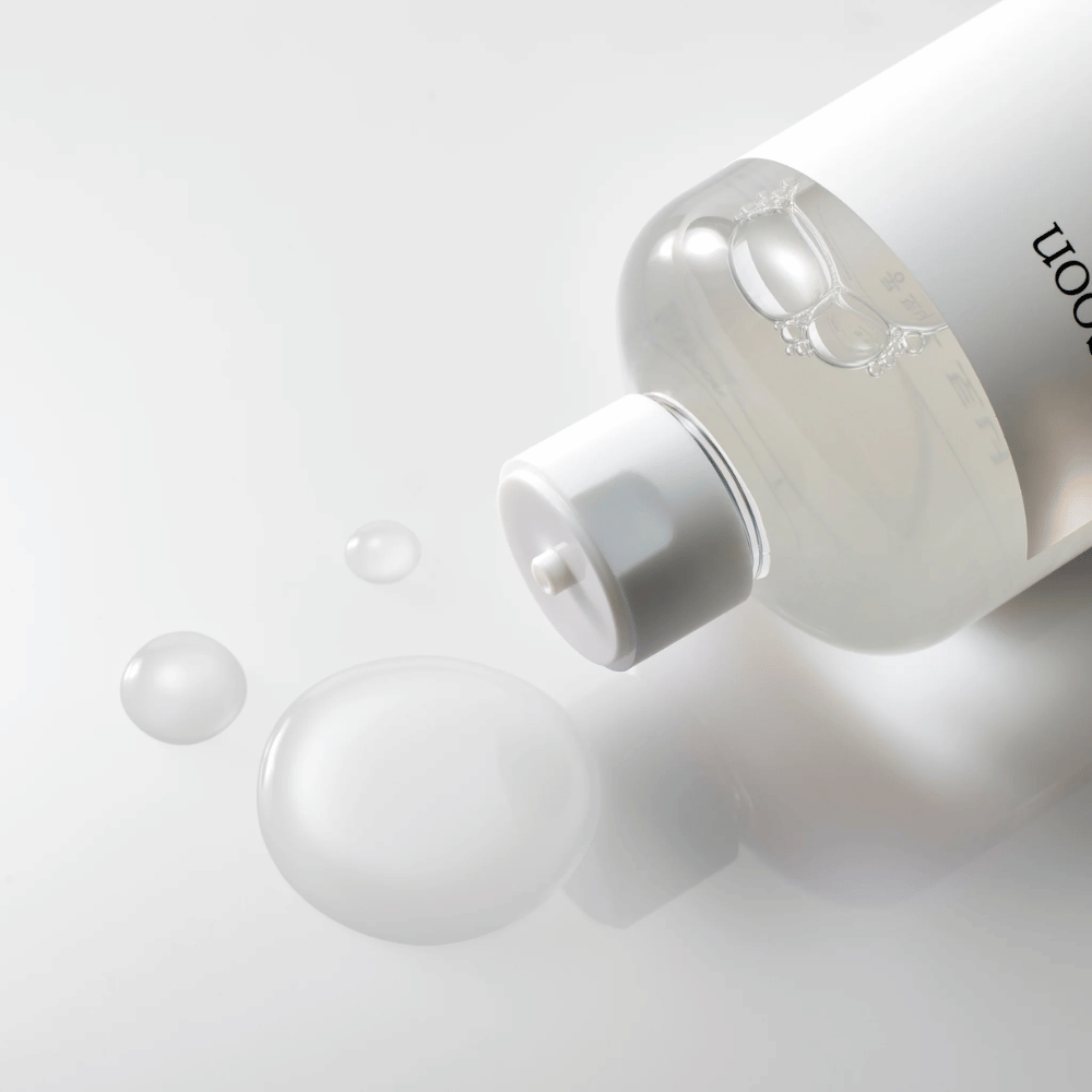 Uma garrafa transparente está deitada de lado sobre uma superfície brilhante com um leve tônico que escorre lentamente formando gotas e uma maior poça na superfície. A garrafa tem um rótulo branco e um mecanismo de bomba branco. A iluminação é suave e cria uma atmosfera limpa e calmante, enquanto a consistência fluida e a clareza do tônico são destacadas pela forma como ele se espalha na superfície.