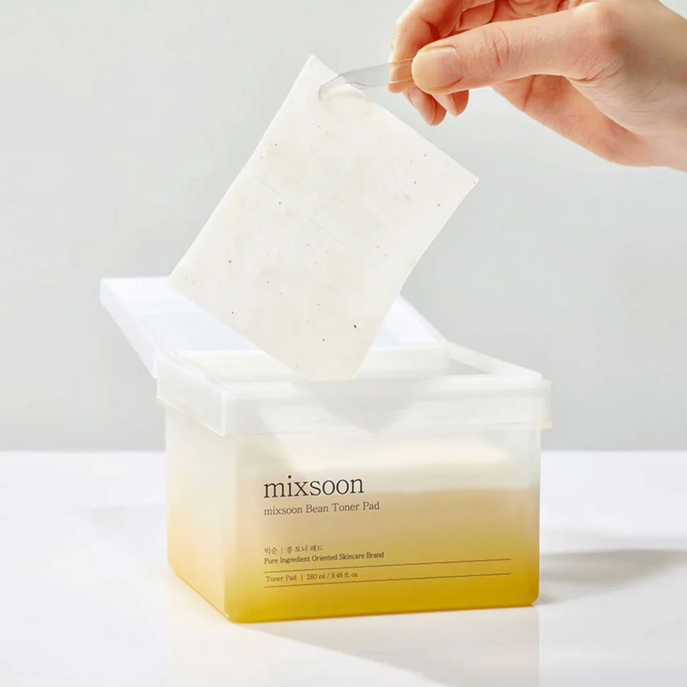 Uma mão segura uma pinça e levanta um Mixsoon Bean Toner Pad de um recipiente quadrado e transparente com um tom amarelado. O recipiente contém 70 pads tonificantes e tem texto que descreve o produto.