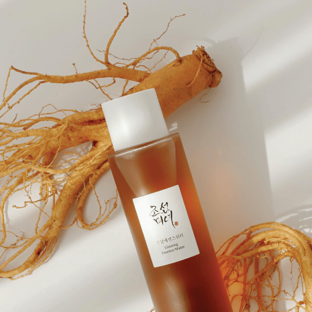 Uma garrafa castanha Beauty of Joseon Ginseng Essence Water com tampa branca está ao lado de uma raiz fresca de ginseng em fundo branco.