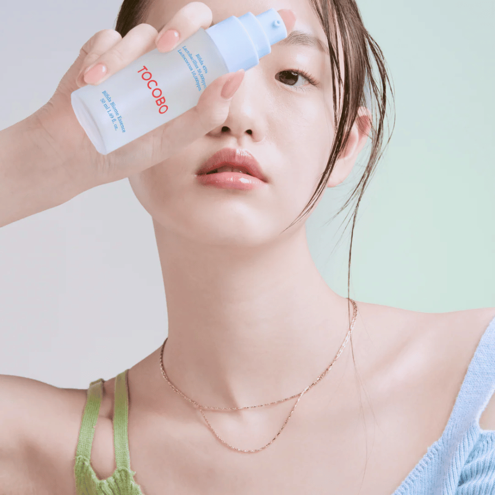Jovem mulher com maquiagem suave e tom de pele natural segura um frasco TOCOBO Bifida Biome Essence em frente ao rosto. Ela usa uma blusa azul-clara com alças verdes e um colar fino de ouro contra um fundo claro.