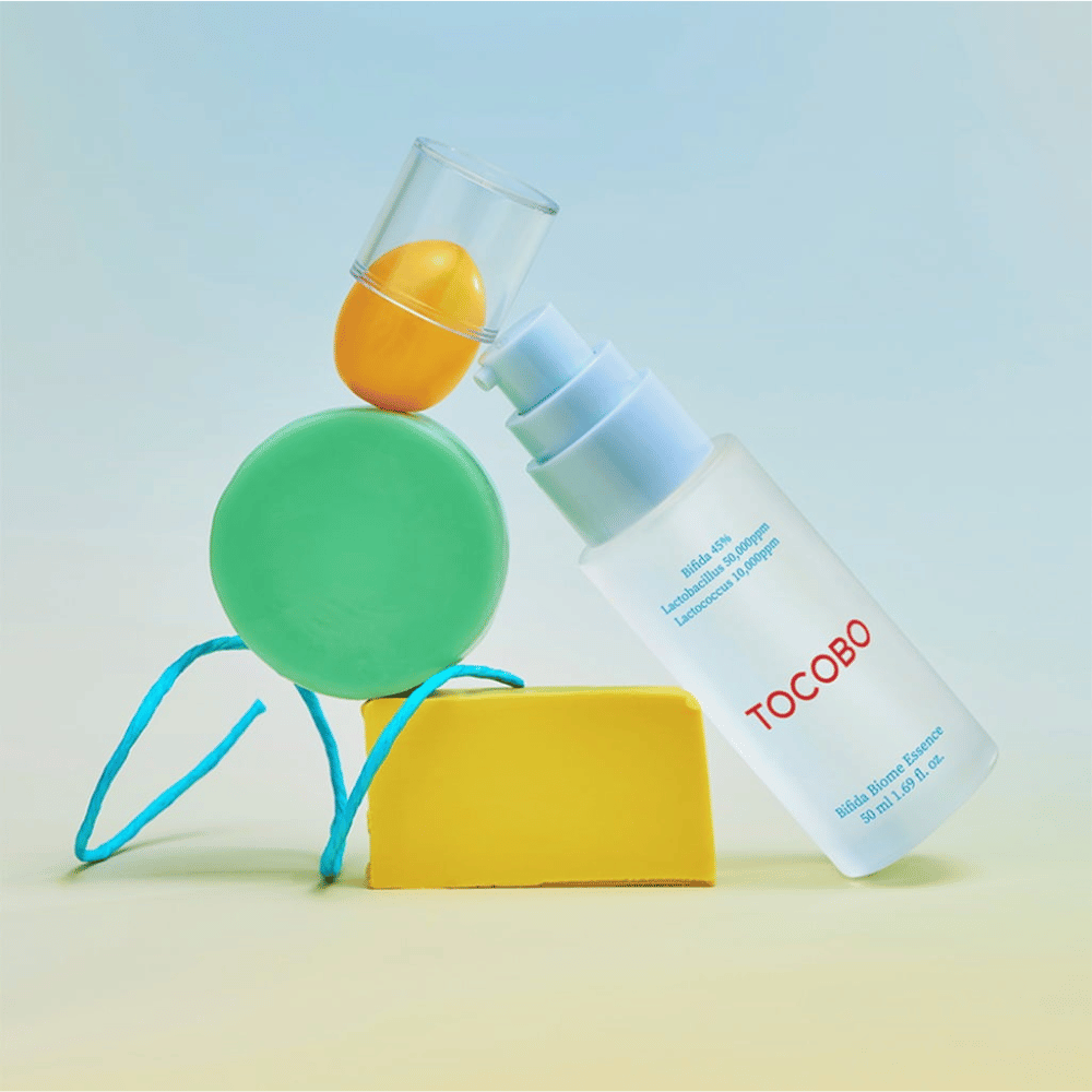 TOCOBO Bifida Biome Essence em um frasco de bomba fosco com logotipo vermelho, colocado contra um fundo colorido com formas geométricas verdes, amarelas e laranja. O texto no frasco indica Bifida 5%, Lactobacillus 3.000 ppm e Lactococcus 1.000 ppm.