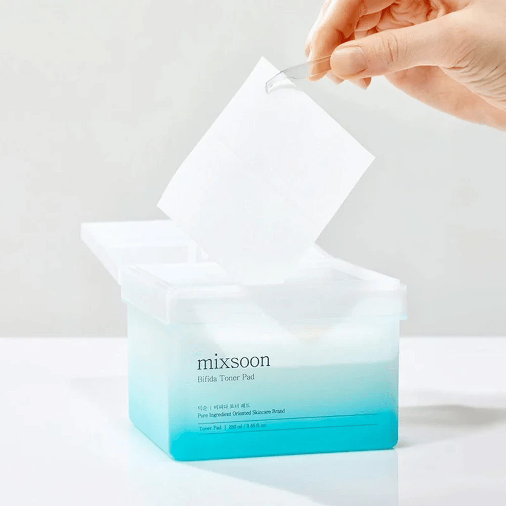 Um recipiente transparente com um gradiente azul e branco que contém discos de toner Bifida. Uma mão retira um disco de toner do recipiente, que está etiquetado com "mixsoon Bifida Toner Pad" e comporta 280 ml (9,46 fl. oz.) de discos de toner destinados aos cuidados com a pele.