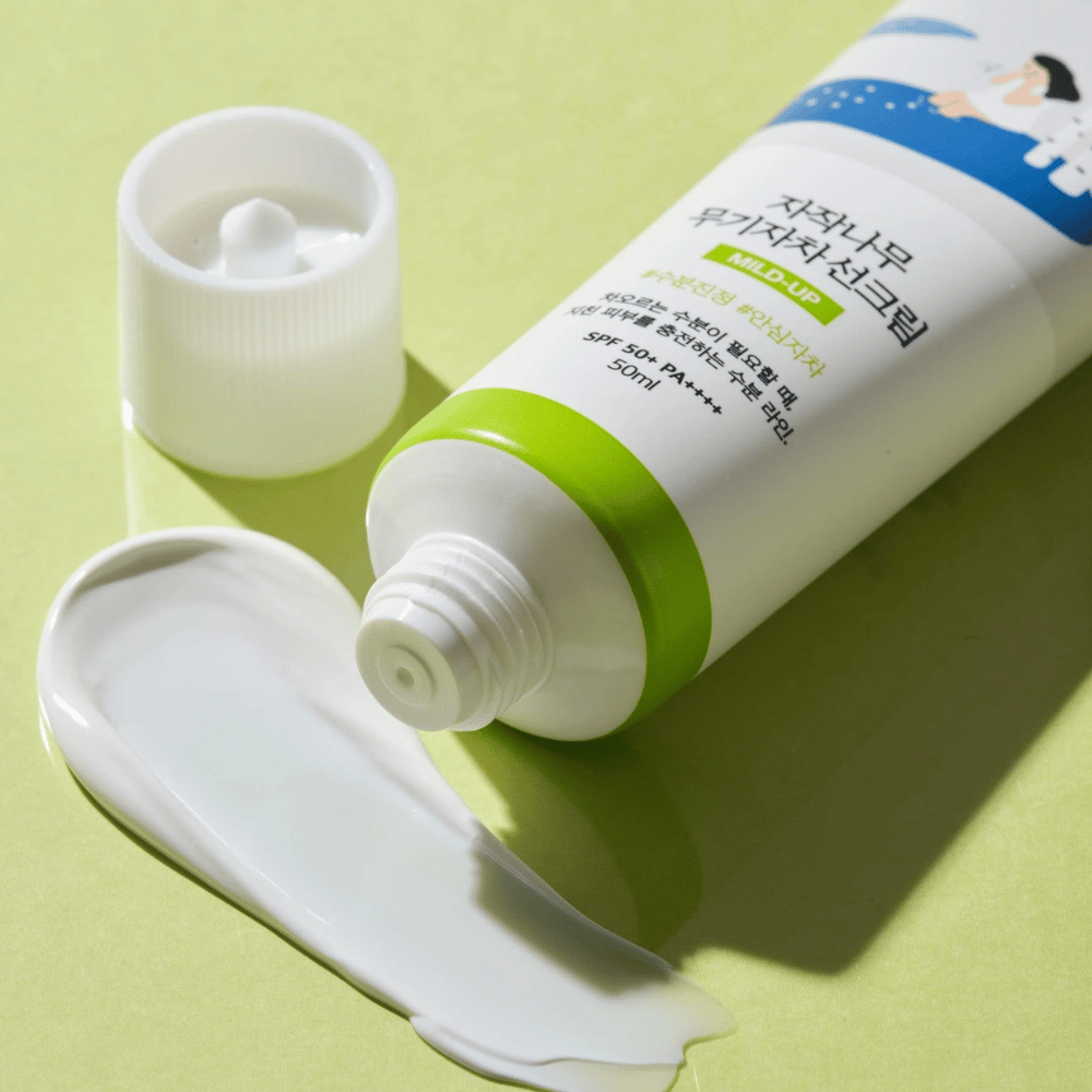 Close-up de Birch Juice Moisturizing Mild Up Sunscreen com tampa desenroscada, creme solar branco espalhado sobre fundo verde claro, SPF 50+ PA++++, 50 ml.