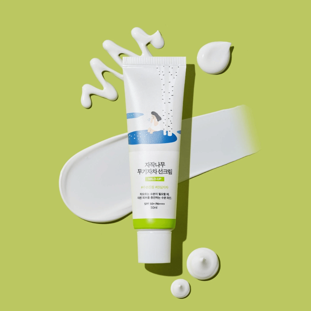 Birch Juice Moisturizing Mild Up Sunscreen – tubo branco com base verde sobre fundo verde, rodeado por creme solar espalhado e gotas, SPF 50+ PA++++, 50 ml.