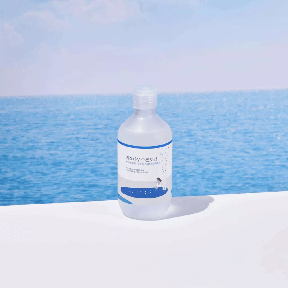 Frasco de ROUND LAB Birch Juice Moisturizing Toner colocado sobre uma superfície branca com mar e céu azul ao fundo