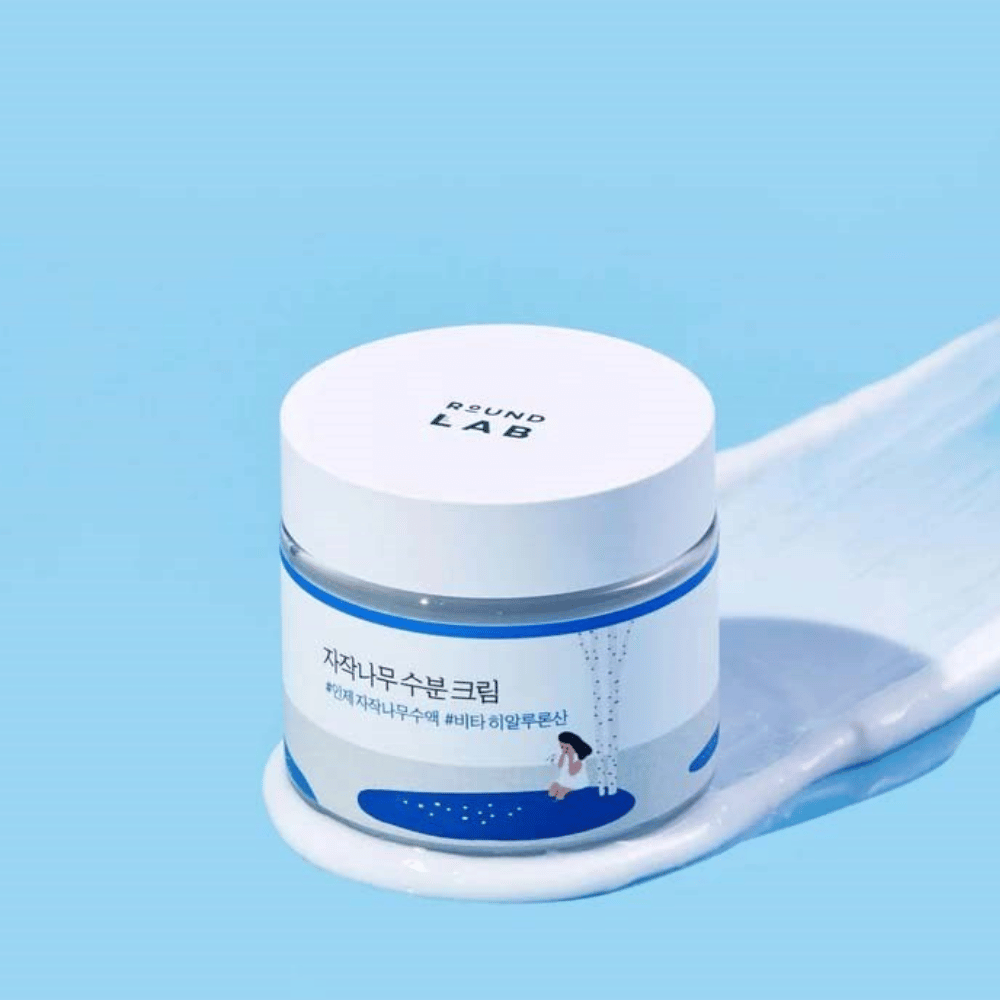 Round Lab Birch Juice Moisturizing Cream em frasco azul com tampa branca, em pé em uma camada de creme contra fundo azul.