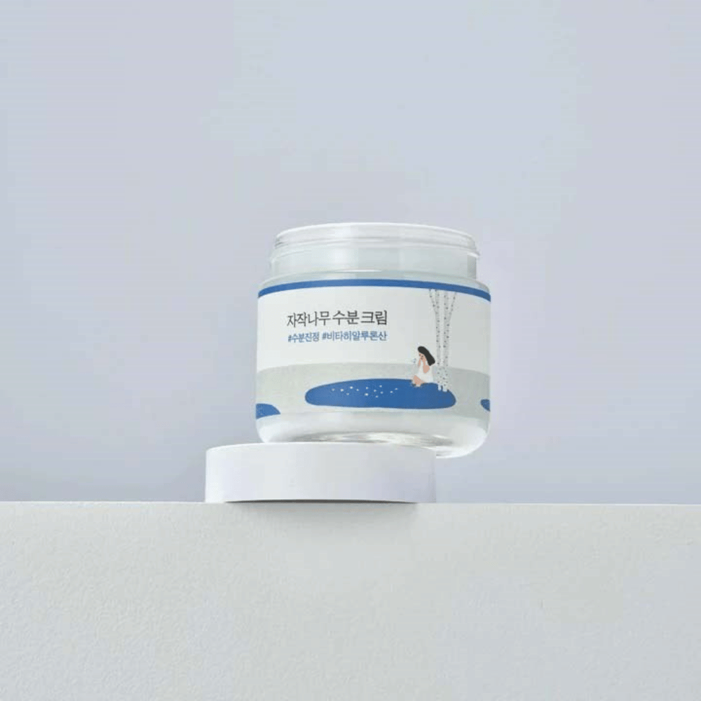Round Lab Birch Juice Moisturizing Cream em frasco transparente com rótulo branco e ilustração azul de mulher junto a bétula, em pé sobre superfície branca contra fundo claro.