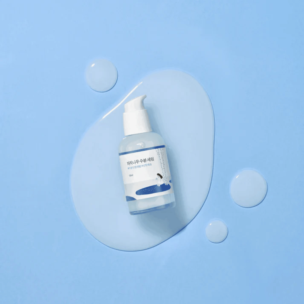 Round Lab Birch Juice Moisturizing Serum em frasco de bomba transparente colocado sobre um fundo azul com gotas e soro derramado.
