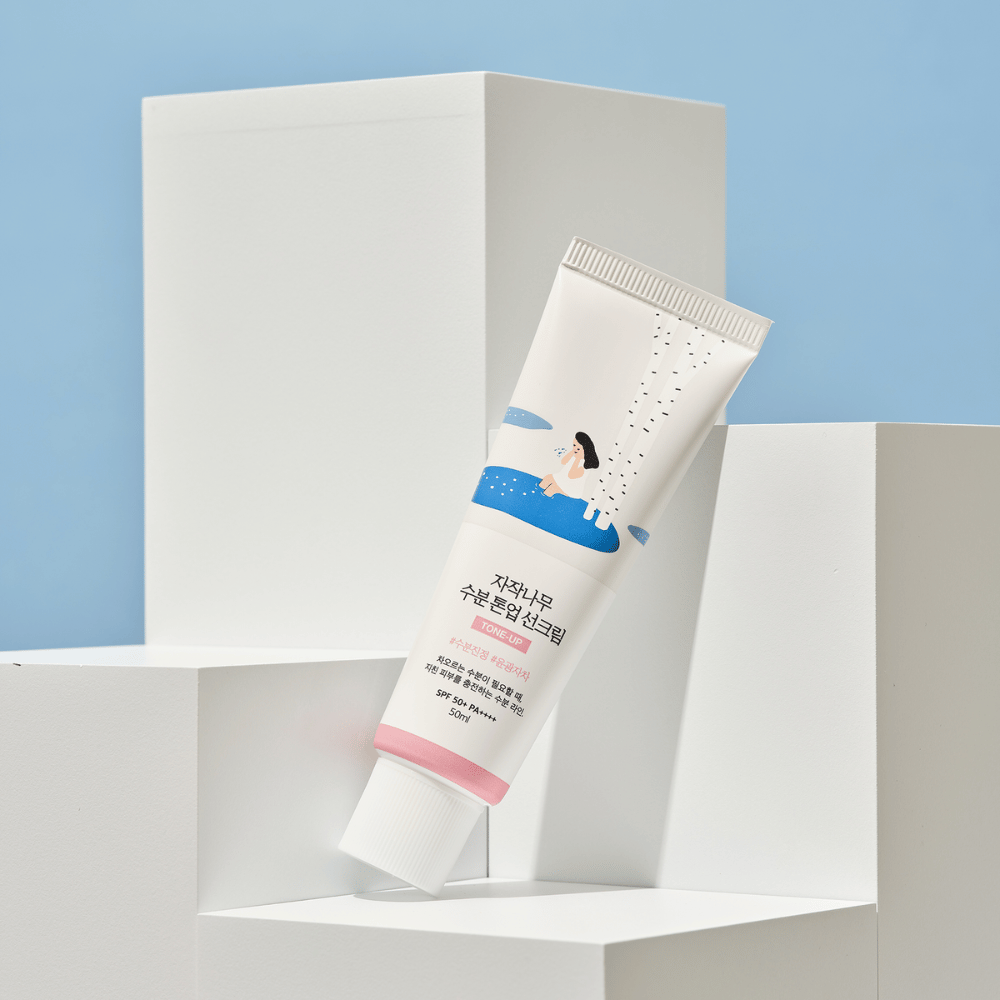Um tubo com Birch Juice Moisturizing Tone Up Sunscreen colocado em cubos brancos contra um fundo azul claro. O tubo é branco com uma ilustração de uma mulher ao lado de uma bétula e água. O texto em coreano indica que o produto tem SPF 50+ PA++++ e contém 50 ml.