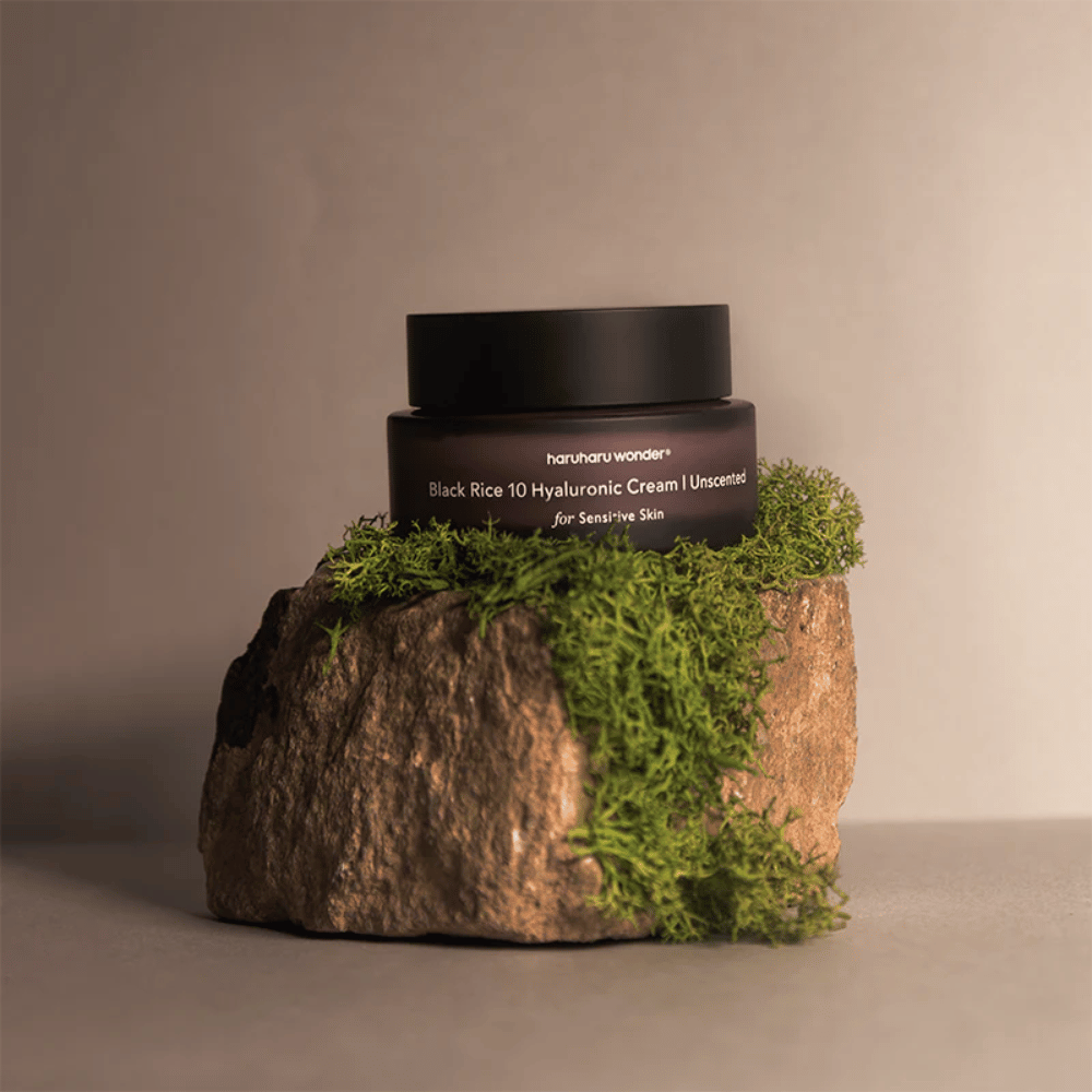 A imagem mostra um pote de creme para a pele marcado "haruharu wonder Black Rice 10 Hyaluronic Cream Unscented for Sensitive Skin". O pote é preto e está sobre uma grande pedra coberta com musgo verde-claro, criando uma sensação natural e terrosa.