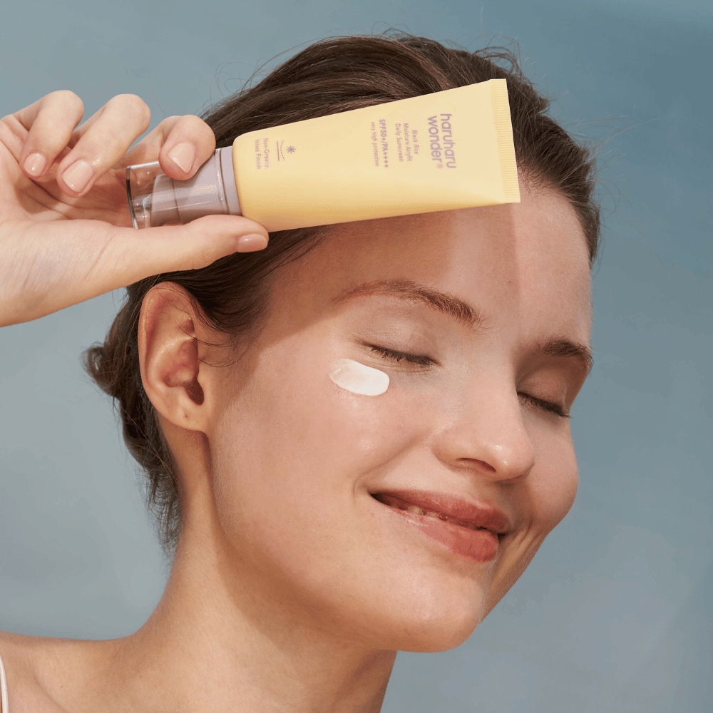 Uma mulher sorridente aplica Black Rice Moisture Airyfit Daily Sunscreen SPF 50+ PA++++ da Haruharu Wonder no rosto. Ela segura um tubo amarelo com protetor solar e tem um pouco do produto na bochecha. O fundo é azul claro.