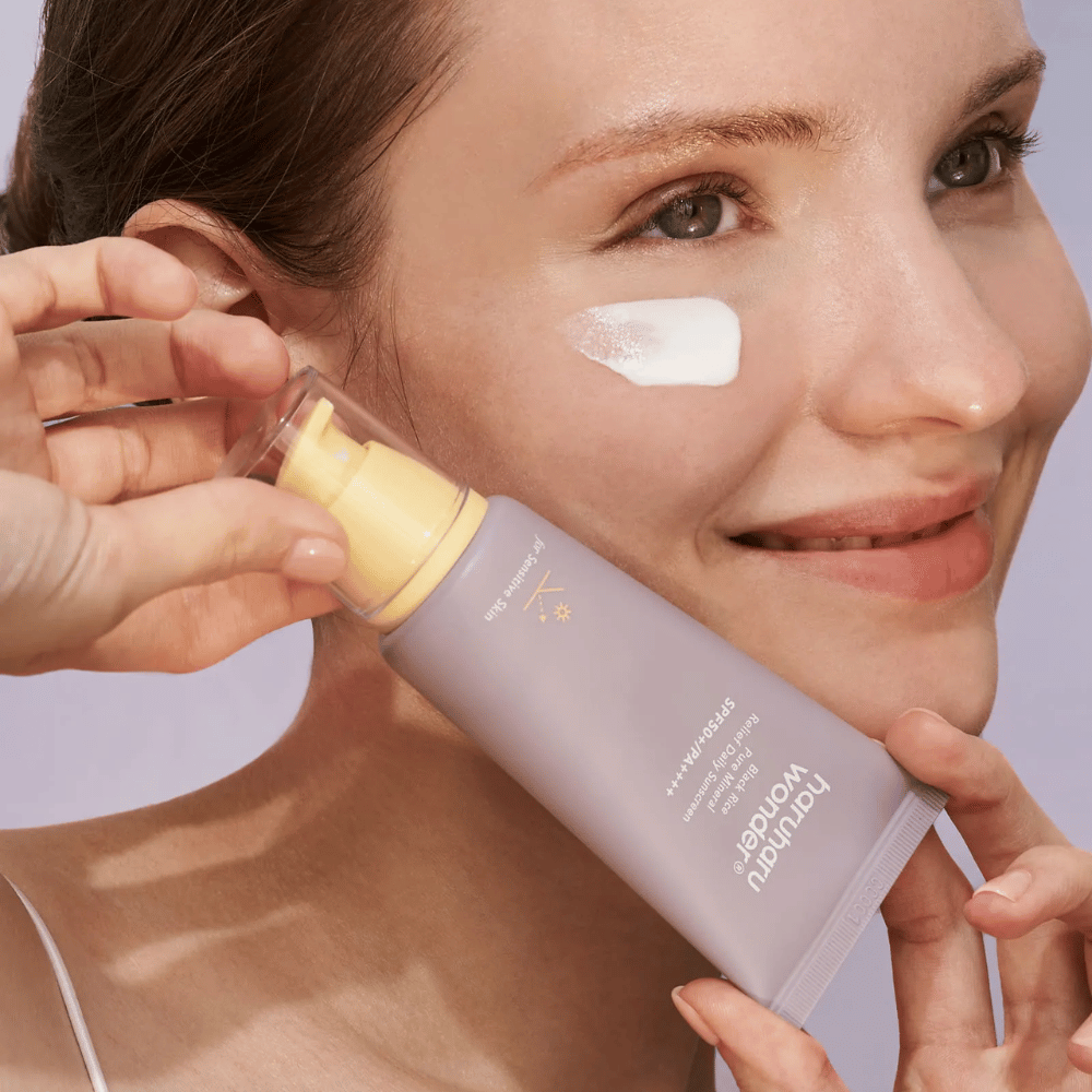 Uma mulher sorridente aplica Haruharu Wonder Black Rice Pure Mineral Relief Daily Sunscreen SPF 50+ PA++++ no rosto. Ela segura um tubo lilás de protetor solar e tem uma gota do produto na bochecha. O fundo é lilás claro.
