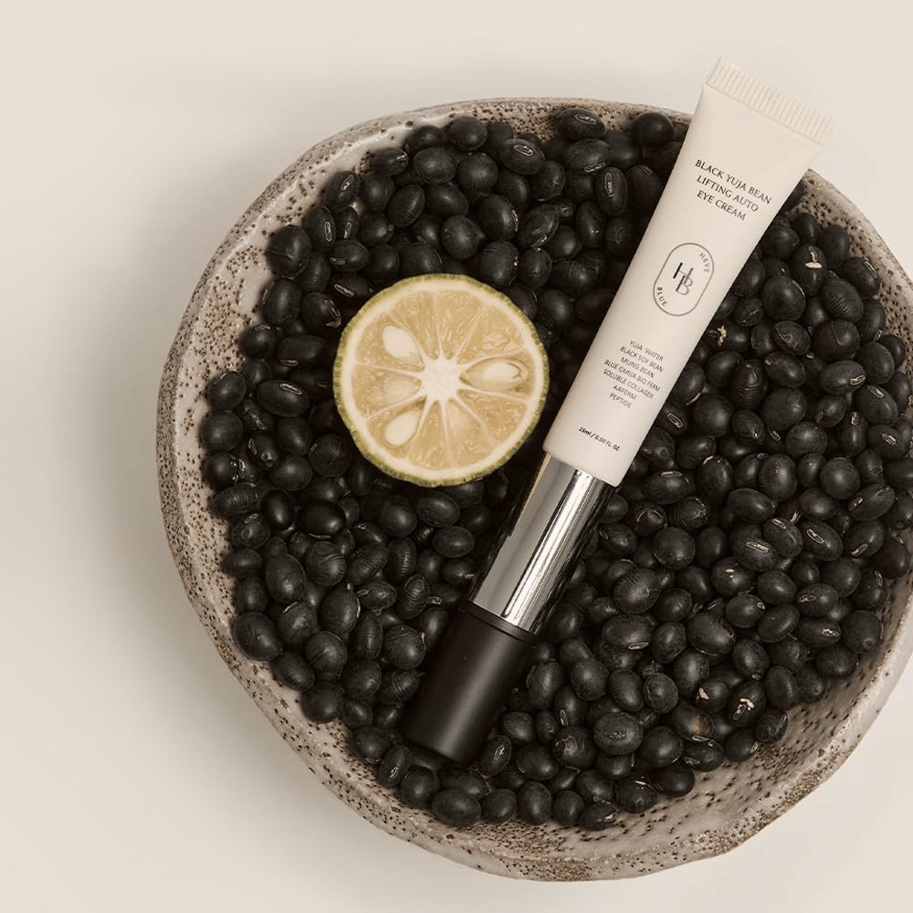 Black Yuja Bean Lifting Auto Eye Cream deitado em uma tigela cheia de feijões pretos e uma fruta cítrica cortada ao meio – uma representação visual dos ingredientes naturais do produto.
