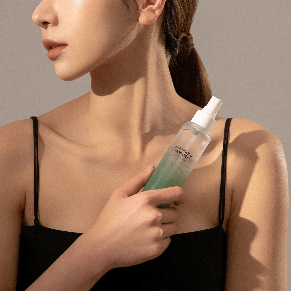 Imagem de HaruHaru Wonder Black Bamboo Mist segurada por uma mulher. O produto está em um frasco de spray transparente com um tom verde e uma bomba branca. A mulher tem um visual descontraído, veste um top preto e está focada em usar a bruma para dar umidade e brilho à pele. A garrafa faz parte de uma rotina de cuidados com a pele.